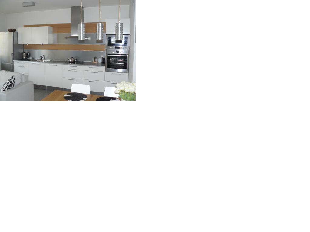 Nowoczesna, jasna kuchnia z białymi meblami, srebrnymi akcentami i drewnianym stołem jadalnym z białymi krzesłami. Nad stołem wiszą trzy cylindryczne lampy. Na stole bukiet białych tulipanów.